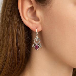 May include: Silver-toned dangle earring featuring a heart-shaped design with ornate detailing. The earring has a central oval-shaped, deep red gemstone. The earring is suspended from a simple hook.