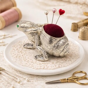 Op de afbeelding: Een zilveren kikker-vormige speldenkussen met een dieprood kussen en drie spelden met parel- en hartvormige koppen. De kikker staat op een wit bord met een kanten placemat, omringd door naaiartikelen zoals vingerhoeden en scharen.