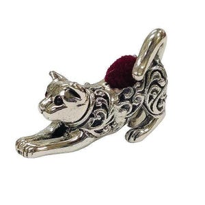 May include: A silver-colored cat-shaped pin cushion with a burgundy cushion. The cat is in a stretching pose, with intricate scrollwork detailing its body. The pin cushion is a decorative sewing accessory.