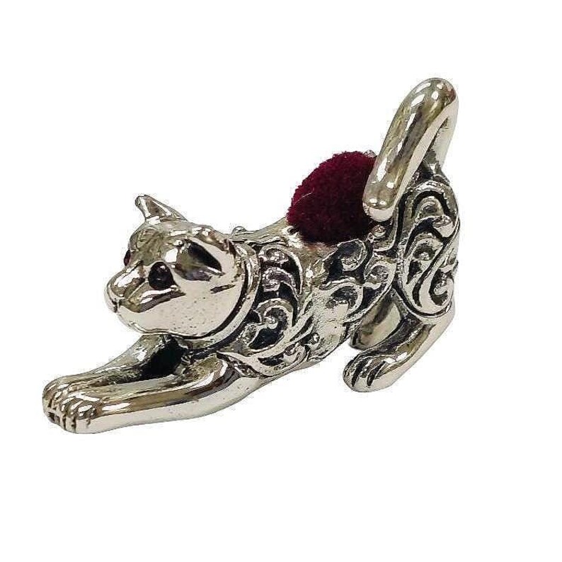 May include: A silver-colored cat-shaped pin cushion with a burgundy cushion. The cat is in a stretching pose, with intricate scrollwork detailing its body. The pin cushion is a decorative sewing accessory.