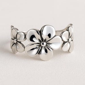 Könnte beinhalten: Ein silberfarbener Ring mit drei stilisierten Blumenmotiven. Jede Blume hat abgerundete Blütenblätter und ein zentrales Detail. Das Design des Rings bildet ein Blumenband. Vor neutralem Hintergrund.