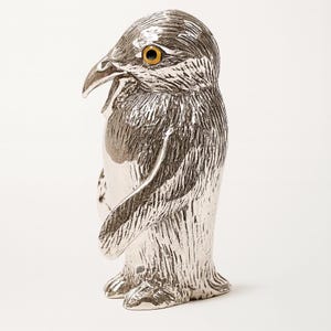 May include: A silver-colored penguin figurine with textured details. The penguin has a detailed beak, yellow eyes, and a white chest. The figurine is standing upright, showcasing its feet and wings. The overall design is detailed and artistic.