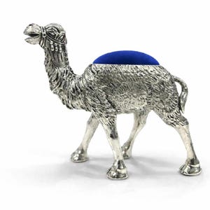May include: A silver-colored camel figurine with a blue cushion on its back. The camel has detailed fur texture and decorative headwear. The figurine is standing on four legs, with its mouth slightly open. A unique decorative item.