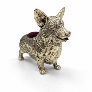 May include: A silver-toned corgi ornament with a textured surface. The dog has a small opening on its back, lined with a deep red material. The figurine is standing on a white surface. The corgi has upright ears.