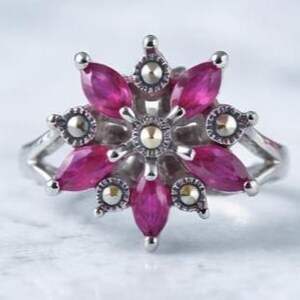 May include: A silver-toned ring featuring a floral design with marquise-cut, deep pink gemstones. The petals are arranged around a central gold-toned accent, with smaller silver and gold details. The ring has a split shank.