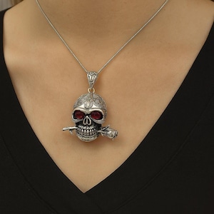 Puede incluir: Un collar con colgante de calavera plateada con ojos de piedras preciosas rojas y una rosa en la boca. La calavera tiene detalles intrincados en la frente y está suspendida de una delicada cadena de plata. Una joya única.