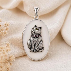 May include: Silver oval pendant with a detailed cat design. The cat has green eyes and a collar with red accents. The pendant hangs from a silver bail and is set against a white background.