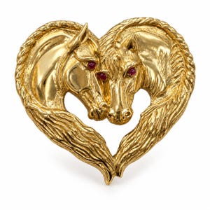 May include: A gold-toned metal brooch shaped like a heart, featuring two horse heads facing each other. The horses have detailed manes and small, round, dark red stones set as eyes. The metal surface has a polished, reflective finish throughout.