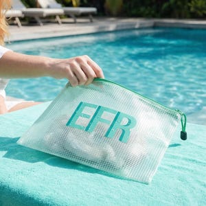 Op de afbeelding: Een witte mesh-ritszak met de letters "EFR" in turquoise. De tas heeft een groene rits en ligt op een turquoise handdoek bij een zwembad. De achtergrond omvat een persoon en een zwembad.