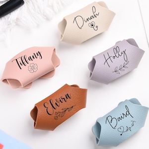 May include: Five colourful leather name tags in pink, beige, lilac, brown, and blue. Each tag has a different name in cursive script with a small decorative element, such as a flower or heart symbol.