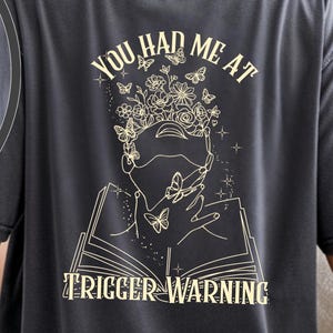 May include: Dark gray t-shirt with a front and back design. The front features a floral and book design. The back has the text "YOU HAD ME AT" above a graphic of a person with flowers and butterflies, and the text "TRIGGER WARNING" below.