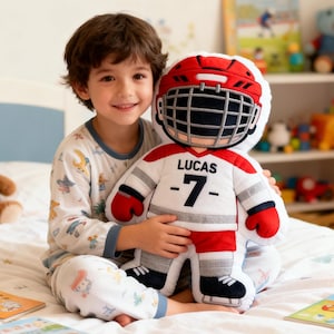 May include: A child in pyjamas smiles, holding a hockey player plush toy. The toy is white, red, and grey, with the name "LUCAS" and the number "-7-" on the front. A red helmet with a black cage covers the head of the plush.