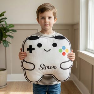 May include: A white, plush video game controller-shaped pillow with black trim and details. The controller features a directional pad, colorful buttons, and the name "Simon" in black script. The pillow is being held by a person.