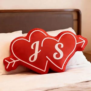 Op de afbeelding: Rood hartvormig kussen met witte omtrek en pijldesign. Het kussen heeft de letters "J" en "S" in witte letters. Het kussen ligt op een wit bed met een bruin hoofdbord op de achtergrond.