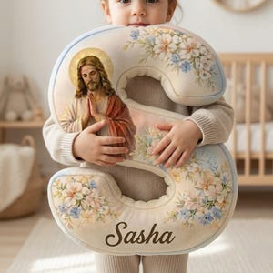 Op de afbeelding: Een crèmekleurig, pluche letter "S" kussen met bloemaccenten en een portret van Jezus. Het kussen heeft een lichtblauwe rand en de naam "Sasha" in een bruin script.