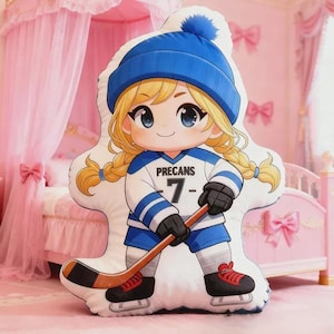 May include: A plush pillow in the shape of a cartoon girl hockey player. She wears a blue beanie, a white and blue jersey with the number 7, black gloves, and skates. The pillow has blonde pigtails and holds a hockey stick.