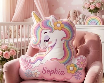May include: A plush unicorn pillow with a rainbow mane and a golden horn rests on a pink armchair. The pillow has the name "Sophia" on a pink cloud. The room has a pink theme with a crib and floral accents.