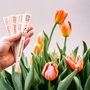 May include: A hand holds a wooden fan with a laser-cut design and the text "Amanda & Drew 22.06.24". The fan is in front of a bouquet of orange and pink tulips.