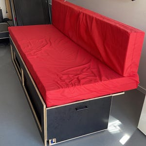 Könnte beinhalten: Ein rotes, gepolstertes Futon mit einem schwarzen Holzrahmen und Staufächern. Das Futon ist ausgeklappt und hat ein rotes Kissen obenauf.