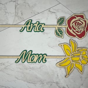May include: Two wooden flower stick decorations with names "Aria" and "Mom" written in green. The flower sticks are on a white marble background. One flower is a red rose and the other is a yellow daffodil.