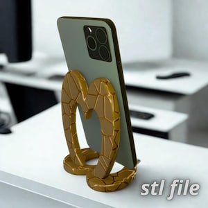 May include: A gold-colored phone stand with a textured, heart-shaped design, holding a green smartphone. The stand is on a white surface, with the text "stl file" in the bottom right corner.