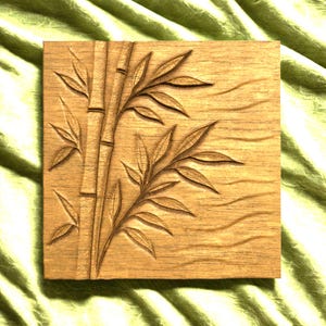 Pode incluir: Placa quadrada de madeira com um desenho de bambu esculpido. Os caules e folhas de bambu são intrincadamente detalhados, criando um efeito tridimensional. A madeira tem um tom quente e natural.