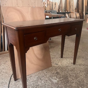 Puede incluir: Un escritorio de madera marrón oscuro con tres cajones y patas delgadas. El escritorio tiene una parte superior rectangular y tiradores de cajón de color latón. El escritorio está en un taller.