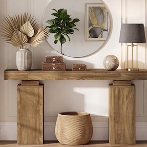 Peut inclure: Une console en bois avec deux pieds rectangulaires épais, comprenant un vase cannelé avec des feuilles de palmier séchées, trois boîtes en bois sculpté, un objet sphérique en pierre, une lampe avec un abat-jour gris et un panier tressé.
