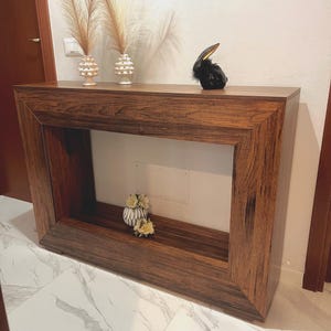 Puede incluir: Una consola rectangular marrón oscuro con un diseño enmarcado. La mesa tiene una figura decorativa de conejo negro y dos jarrones con altas plumas. Un pequeño jarrón con flores está dentro de la mesa.