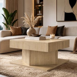 Peut inclure: Une table basse carrée en bois clair avec un socle. La table est dans un salon avec un canapé beige, un vase décoratif et des livres. La pièce a une palette de couleurs neutres avec de la lumière naturelle.