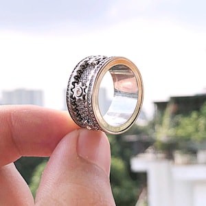 Peut inclure: Une bague argentée avec un design détaillé de rouages et de motifs floraux. La bague a une bande intérieure dorée. La bague est présentée sur fond de ciel nuageux et de bâtiments.
