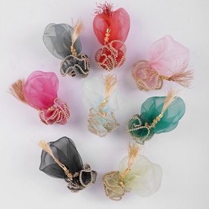 May include: An assortment of decorative fabric ornaments in various colors, including red, pink, green, and gray. Each ornament features a sheer, ruffled base adorned with gold-colored beads and a tassel. The ornaments are arranged on a white surface.