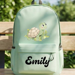 May include: A pale green backpack with a cartoon turtle design. The turtle has a beige shell and is set amongst yellow flowers. The name "Emily" is printed in black script on the front pocket. The backpack has a black handle and zip.