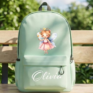 Könnte beinhalten: Hellgrüner Rucksack mit Feen-Grafik. Die Fee hat eine Krone, Flügel und einen Zauberstab. Der Name "Olivia" steht in Schreibschrift auf der Vordertasche. Der Rucksack hat einen schwarzen Griff und Reißverschluss.