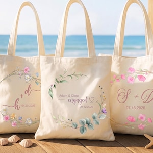 Pode incluir: Três sacolas de lona na cor creme com desenhos de guirlandas florais. A bolsa da esquerda mostra 'h + d' e '04.20.2026', a central 'Adam & Clara engaged' e '09.12.2026', e a da direita 'S + D' e '07.16.2026' em letras rosas.