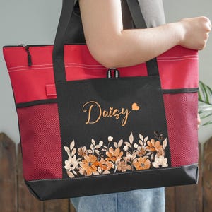 May include: A red and black tote bag with a floral design and the name "Daisy" in cursive. The bag has a zippered top, mesh side pockets, and a black handle. The floral design features orange and white flowers.