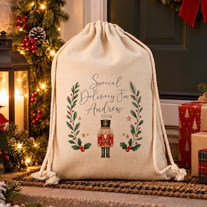 May include: A beige drawstring canvas sack featuring the printed text Special Delivery For Andrew 2026, flanked by two green leafy branches with red berries and gold stars, and a central illustration of a red and green nutcracker.