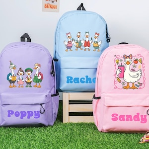 Op de afbeelding: Drie kleurrijke rugzakken met cartoon ganzen ontwerpen. Een lavendelkleurige rugzak heeft de naam "Poppy" in paars. Een lichtblauwe rugzak heeft de naam "Rachel" in blauw. Een roze rugzak heeft de naam "Sandy" in roze.
