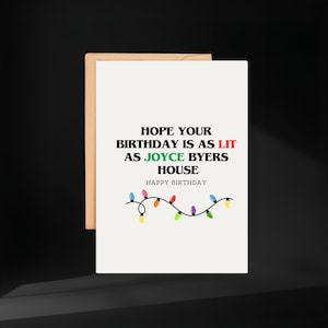 Può includere: Un biglietto di auguri bianco con la scritta "HOPE YOUR BIRTHDAY IS AS LIT AS JOYCE BYERS HOUSE HAPPY BIRTHDAY" in nero, rosso e verde. Una fila di luci colorate decora il fondo.