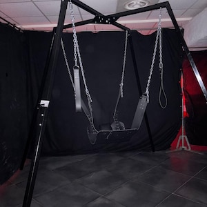 Könnte beinhalten: Ein schwarzes Metall-Schaukelgestell mit einem schwarzen Lederschaukelsitz und silbernen Ketten. Das Schaukelgestell steht in einem Raum mit schwarzen Vorhängen und einem gefliesten Boden.