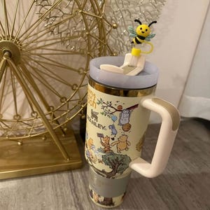May include: A Stanley travel mug featuring Winnie the Pooh characters. The mug has a light yellow background with a gold rim and a white and tan handle. A bee-shaped lid topper adds a whimsical touch.