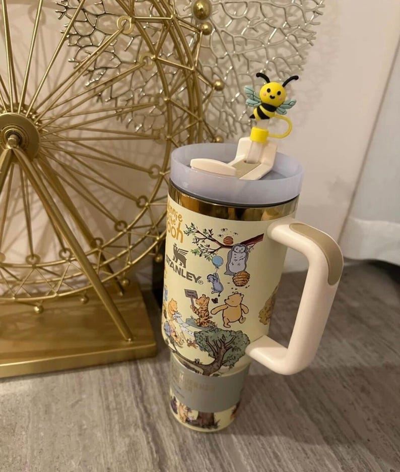 May include: A Stanley travel mug featuring Winnie the Pooh characters. The mug has a light yellow background with a gold rim and a white and tan handle. A bee-shaped lid topper adds a whimsical touch.