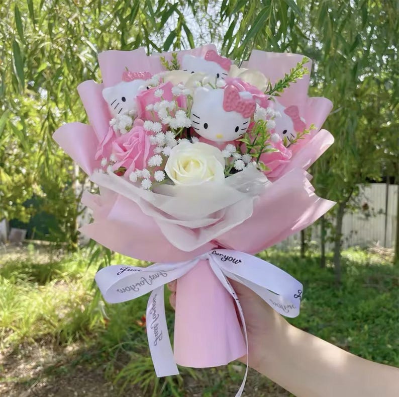 May include: A bouquet featuring pink roses, white roses, and Hello Kitty figures. The arrangement is wrapped in pink paper and tied with a white ribbon that has text on it. The bouquet is held in a person's hand.