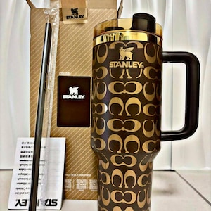 Puede incluir: Un vaso Stanley marrón y dorado con asa, con un patrón repetido. El vaso tiene una tapa y un asa negras, y un borde dorado. También son visibles una pajita negra y el embalaje, con el logotipo de Stanley.