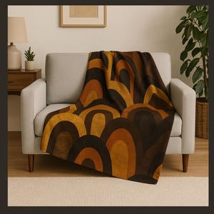 Könnte beinhalten: Eine Decke mit einem Muster aus braunen, beigen und orangefarbenen Bögen, die über einem grauen Sofa drapiert ist. Das Sofa steht vor einer weißen Wand mit einem gerahmten Bild.