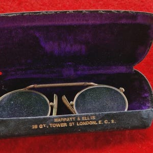 Könnte beinhalten: Antike runde Brille mit goldfarbenem Steg und Rahmen, in einem offenen, dunklen Etui mit lila Samtfutter. Das Etui ist schwarz mit goldfarbener Schrift: "MARRATT & ELLIS 28 GT. TOWER ST LONDON. E.C.S."
