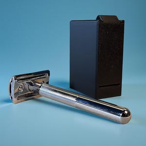 Könnte beinhalten: Ein verchromter Rasierer mit geriffeltem Griff liegt neben einem schwarzen Rasiererständer. Der Rasierer hat ein klassisches Design mit silbernem Finish. Der Ständer ist rechteckig und hat eine strukturierte Vorderseite.