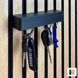 Könnte beinhalten: Ein schwarzer Schlüsselhalter mit mehreren Schlüsseln, die daran hängen. Der Schlüsselhalter ist an einer Holzwand mit schwarzen vertikalen Streifen befestigt. Ein Schlüssel hat einen Yamaha-Schlüsselanhänger.