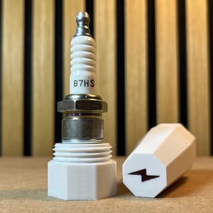 Op de afbeelding: Een bougie met de tekst "B7HS" en een witte, zeshoekige basis. Een witte, zeshoekige dop met een bliksemsymbool staat naast de bougie. De achtergrond is een houten muur.