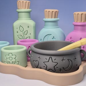 May include: A set of pastel-coloured potion bottles, small cups, and a dark grey cauldron with a yellow stirring tool, all arranged on a beige wavy tray. The bottles feature embossed plant, moon, and star designs, while the cups show flower and star motifs.